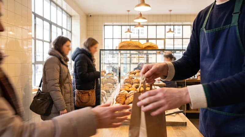 Bäckereitheke: eine Verkäuferin reicht eine Papiertüte über den Tresen, dahinter warten Kunden